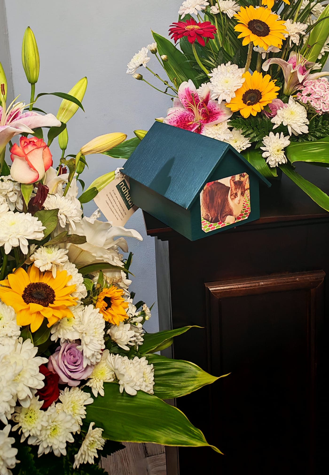 Casita conmemorativa con la foto de una mascota, junto a un arreglo de girasoles y lirios
